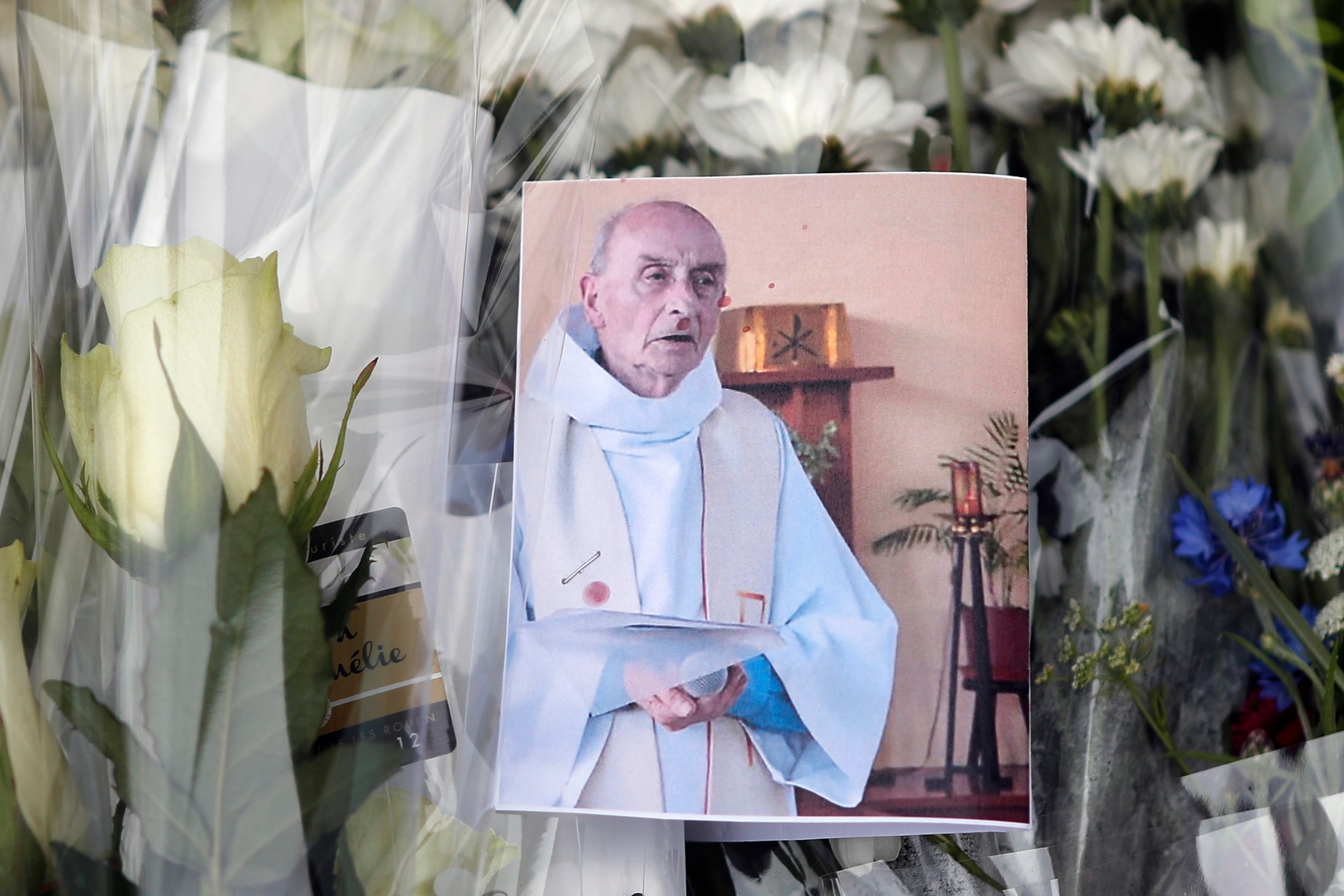 A picture of late Father Jacques Hamel is placed on flowers at the makeshift memorial in front of the city hall closed to the church where an hostage taking left a priest dead the day before in Saint-Etienne-du-Rouvray, Normandy, France, Wednesday, July 27, 2016. The Islamic State group crossed a new threshold Tuesday in its war against the West, as two of its followers targeted a church in Normandy, slitting the throat of an elderly priest celebrating Mass and using hostages as human shields before being shot by police. (AP Photo/Francois Mori) France Hostage Taking