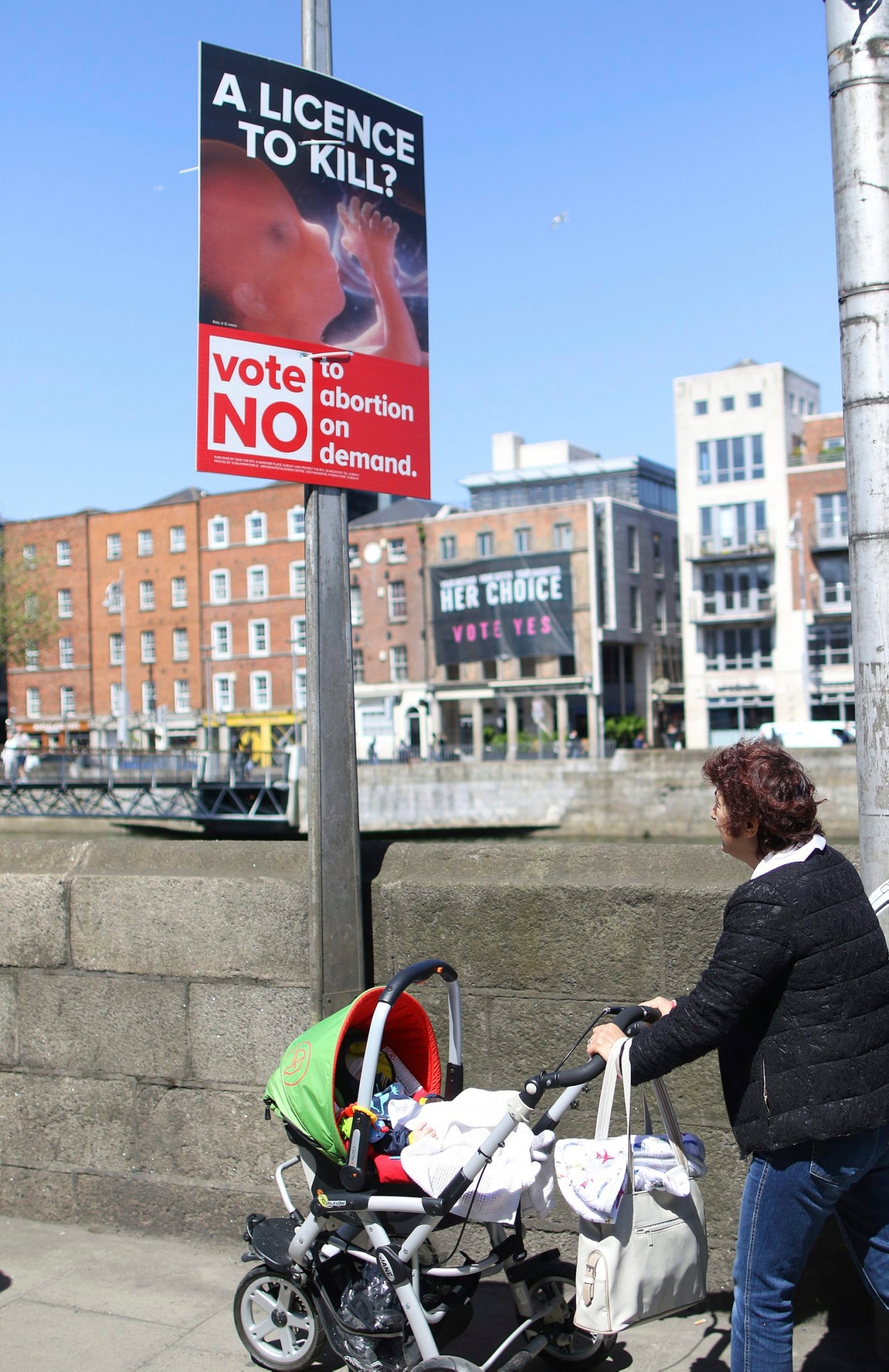 In this photo taken on May 17, 2018,  a woman pushing a pram walks by an anti-abortion poster hung on a lamppost, in Dublin, Ireland. In homes and pubs, on leaflets and lampposts, debate rages in Ireland over whether to lift the country's decades-old ban on abortion. Pro-repeal banners declare: "Her choice: vote yes." Anti-abortion placards warn against a "license to kill." Online, the argument is just as charged _ and more shadowy _ as unregulated ads of uncertain origin battle to sway voters ahead of Friday's referendum, which could give Irish women the right to end their pregnancies for the first time.  (AP Photo/Peter Morrison) Ireland Abortion