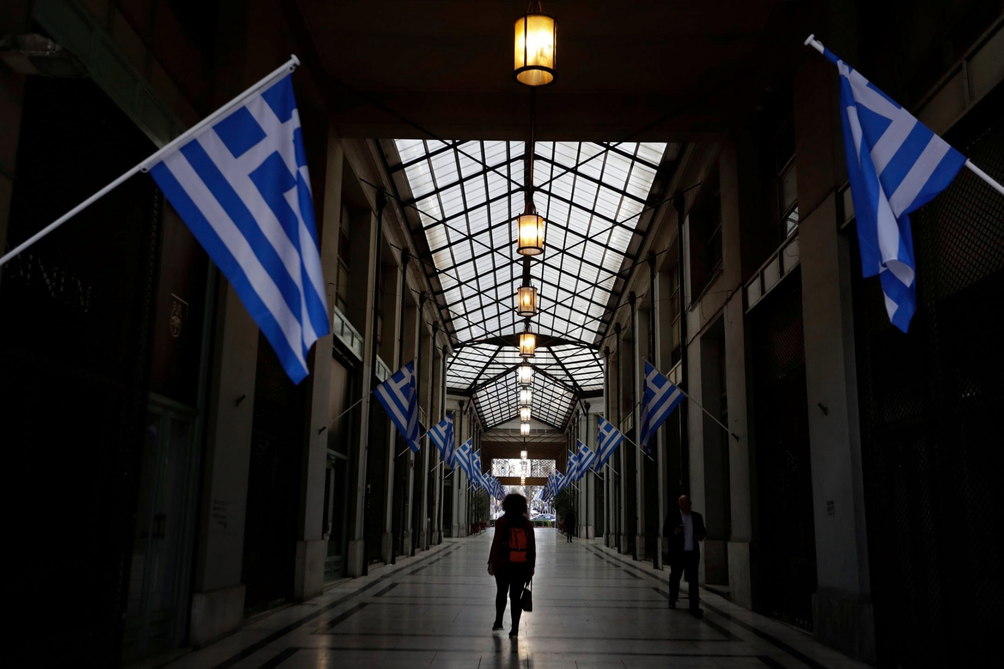In this photo taken on Wednesday, March 22, 2017 a woman passes a shopping arcade with Greek flags, where shops closed because of the crisis in central Athens. Over the past seven years, austerity has left visible scars in GreeceÄôs capital. A walk around Athens reveals more homeless than ever despite some signs of a rosier economic outlook. Thousands of shops, mostly small businesses, are shuttered here and across the country. In what used to be a busy shopping arcade, closed stores are padlocked against a backdrop of hanging Greek flags. (AP Photo/Thanassis Stavrakis) GREECE AUSTERITY PHOTO GALLERY