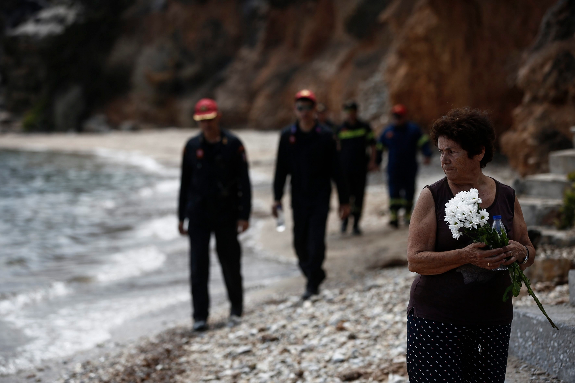 epa06914199 Greek firefighters search for missing people, as Fotini Matrakidis holds flowers next to the point where a six month baby died, following a deadly forest fire in Mati, a northeast suburb of Athens, Greece, 27 July 2018. The confirmed death toll from forest fires that raged through seaside resorts near the Greek capital Athens has increased to 87. According to an announcement by Fire Brigade, the search for more missing persons in the burnt out buildings in eastern Attica is continuing.  EPA/YANNIS KOLESIDIS GREECE WILDFIRE AFTERMATH