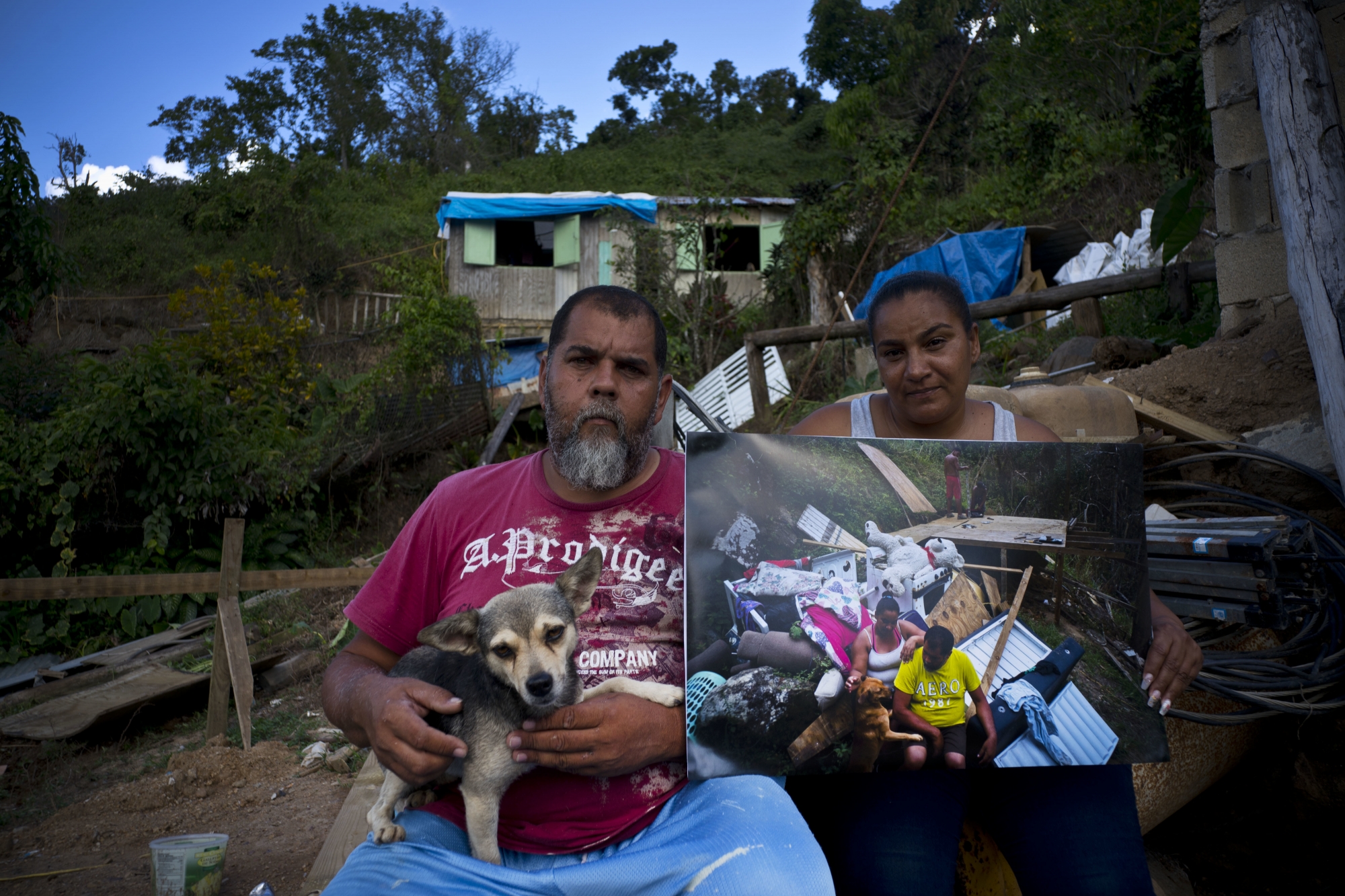 William Fontan Quintero and his wife Yadira Sostre pose with a printed photo of them taken on Sept. 30, 2017 when they sat amid the rubble of their home that was destroyed by Hurricane Maria, at the same spot where they rebuilt their home, behind, in the San Lorenzo neighborhood of Morovis, Puerto Rico, May 29, 2018. The couple says FEMA rejected their application for financial help but they received $8,000 from family to help them replace their belongings, which they invested in wood to build a small home, behind, where they live with their two children who are university students. The roof is plastic tarp while they wait for their FEMA application to be approved so they can finish rebuilding. "We don't have time to build anything safe without help," said Quintero. (AP Photo/Ramon Espinosa) Puerto Rico Hurricane Then and Now Photo Essay